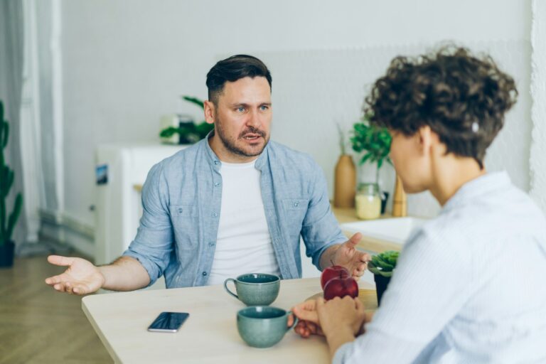 ein Paar, diskutierend am Tisch, er im Fokus, in der aktiven, sprechenden Position, sie undeutlich, von schräg hinten